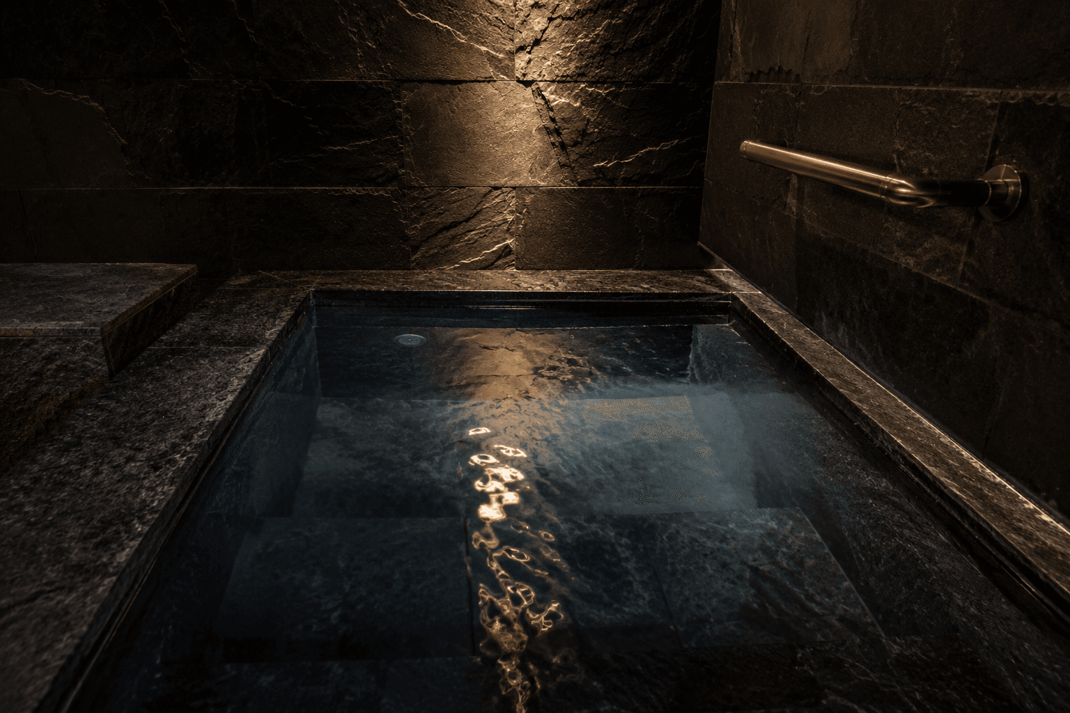The cold plunge at Yumori — a dark stone pool in a basalt room, mineral water under a single warm light, a bronze fixture on the right wall