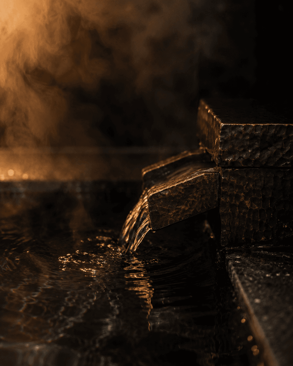 A bronze spout at the edge of a dark mineral onsen at Yumori, water running and steam catching a single warm light against wet basalt