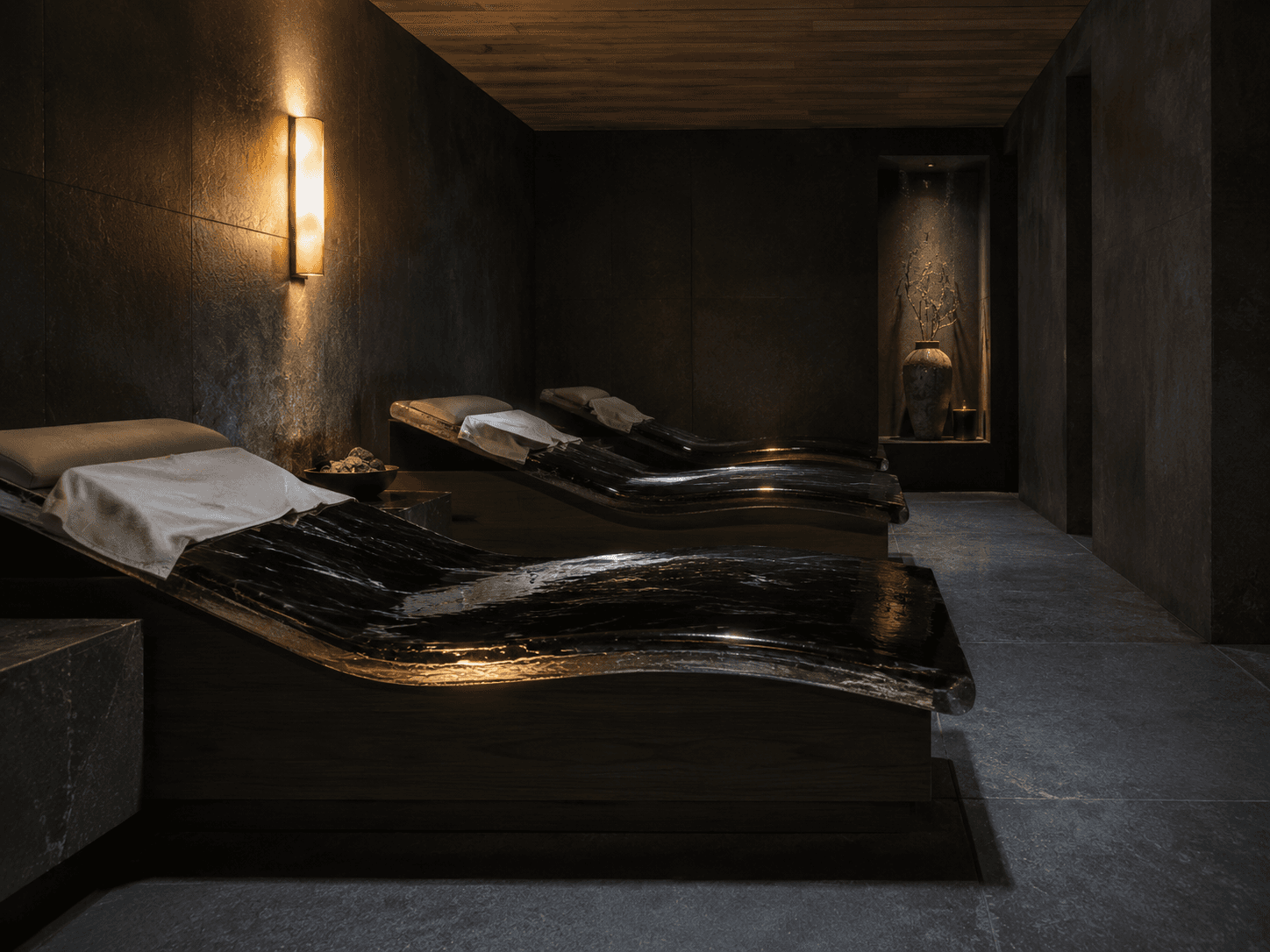 The recline room at Yumori — two heated obsidian loungers draped with white linen, a warm sconce on the left wall, an apothecary vase in a niche to the right