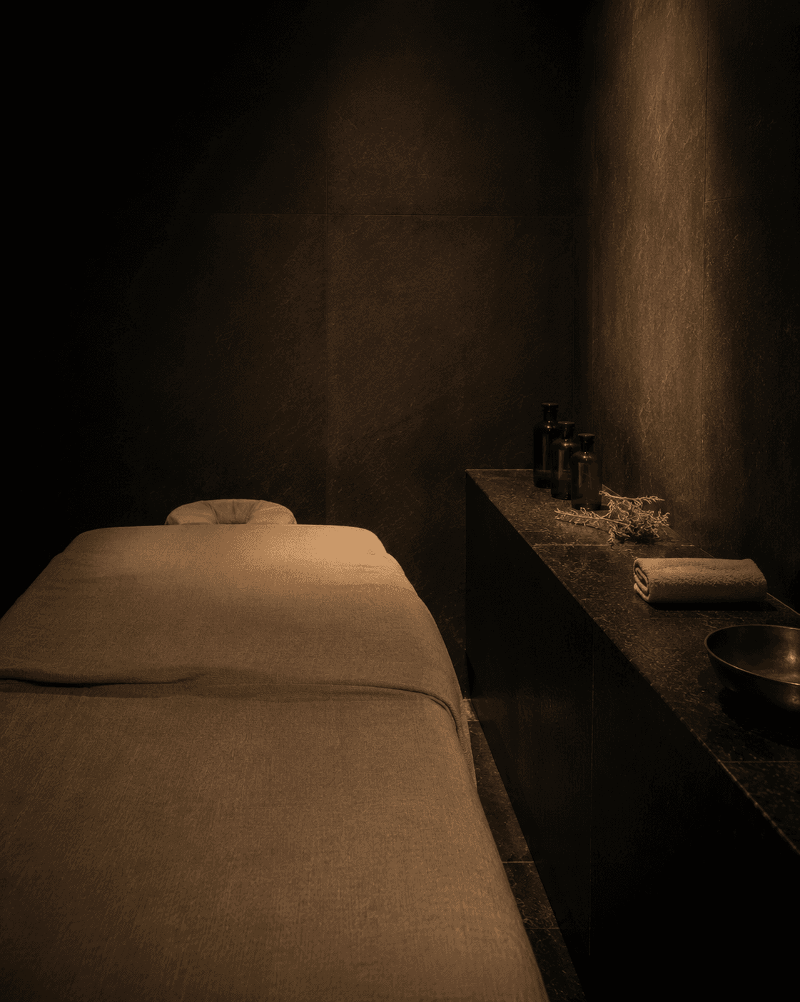 A private treatment room at Yumori — a linen-draped massage table in a dark basalt room, amber oil bottles and a dried botanical sprig on a stone ledge, a bronze bowl